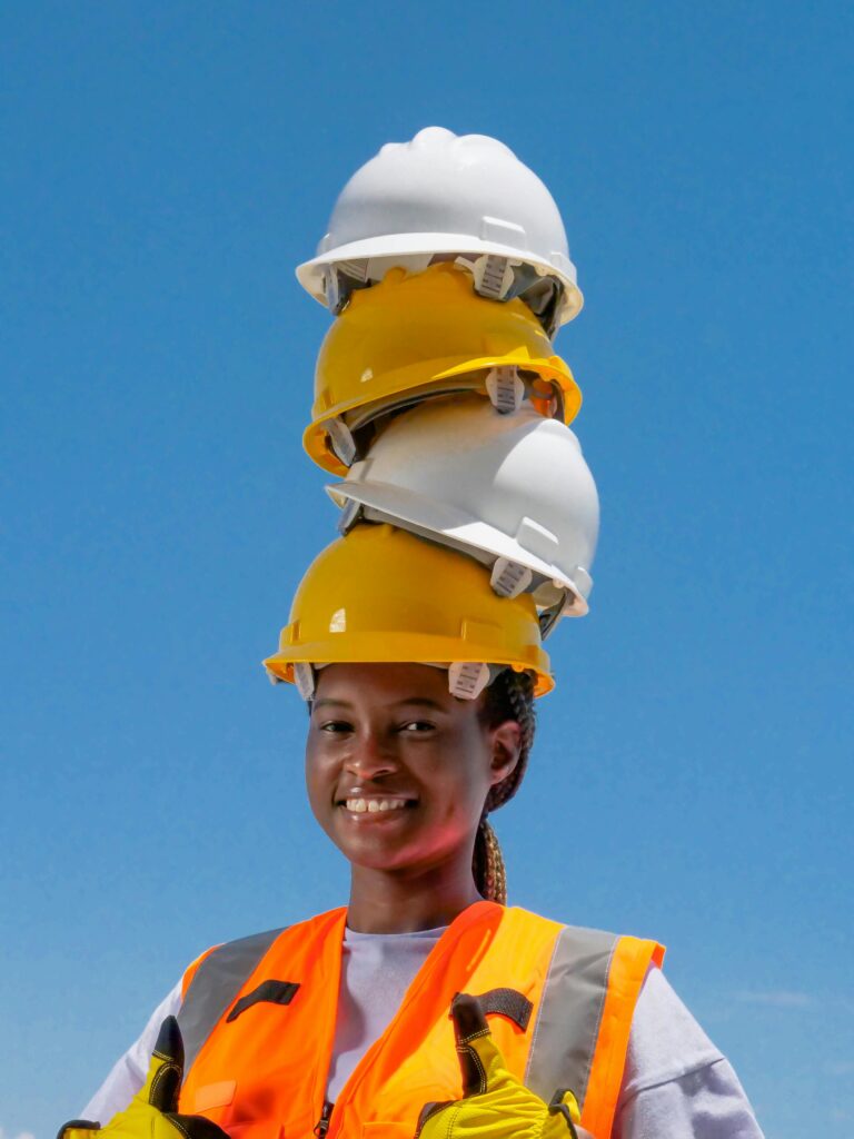 pexels photo 8487770 8487770 Smiling African American woman in safety gear balancing multiple hard hats on head against blue sky.